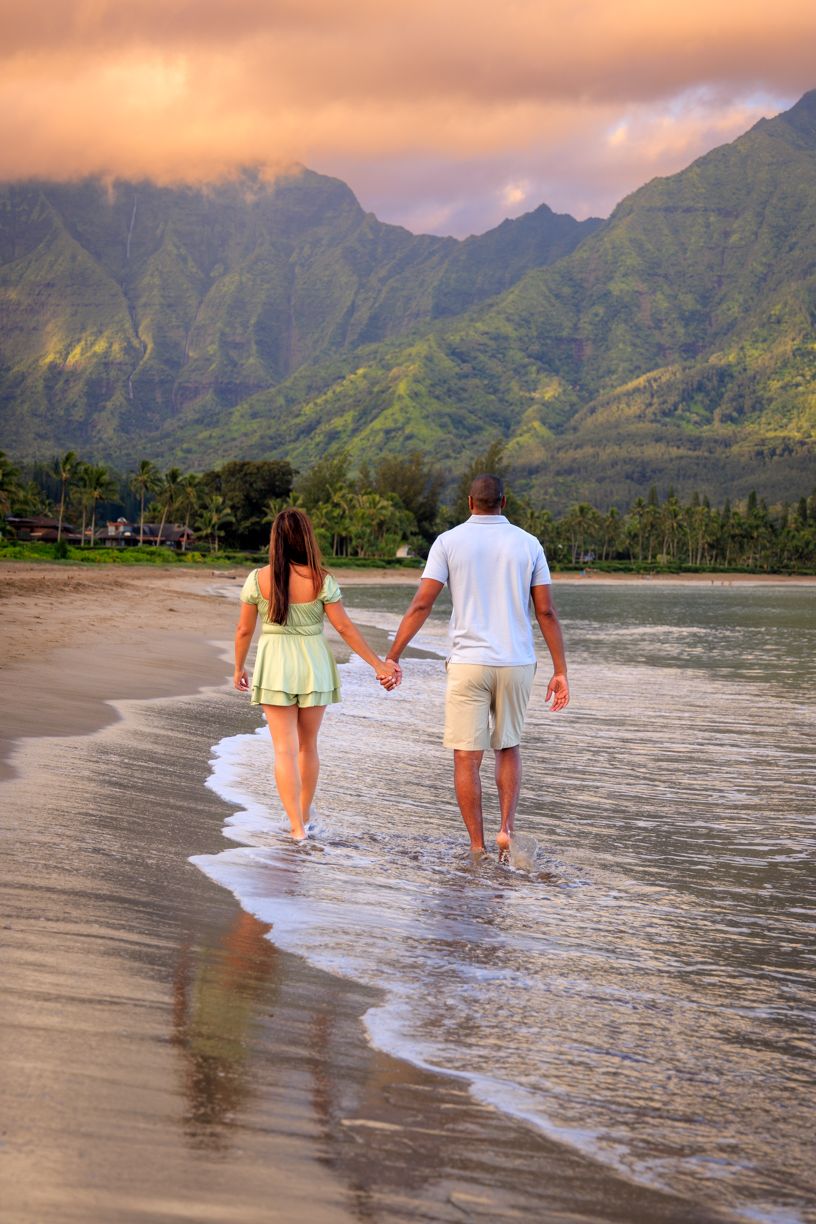 Couple walking along the shoreline at sunset with mountains and palm trees in Hanalei, Hawaii.