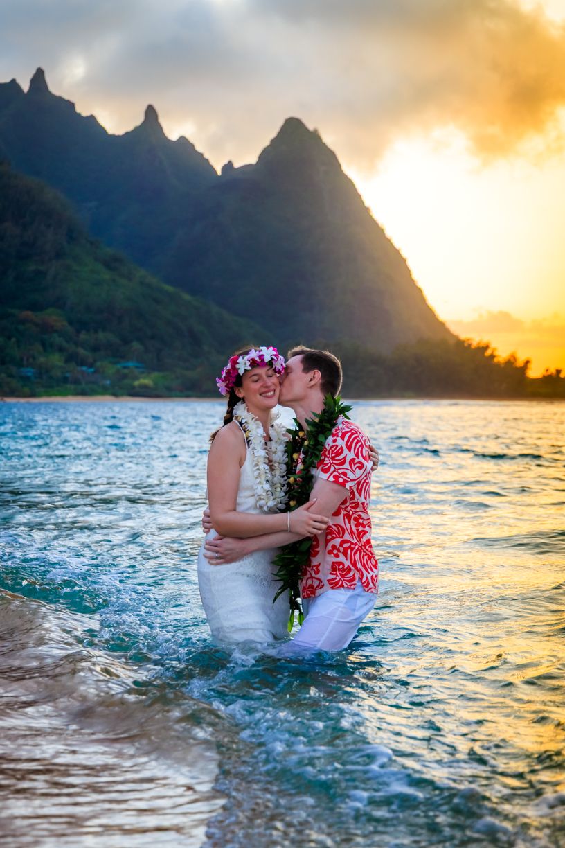 Romantic couple embraces in ocean waters at sunset with dramatic mountain peaks in background.