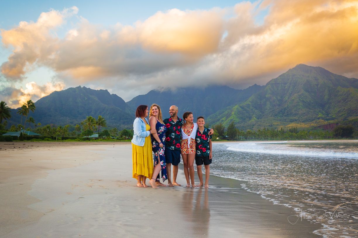 Family group poses together on a scenic Hawaiian beach at sunset with mountains in the background.