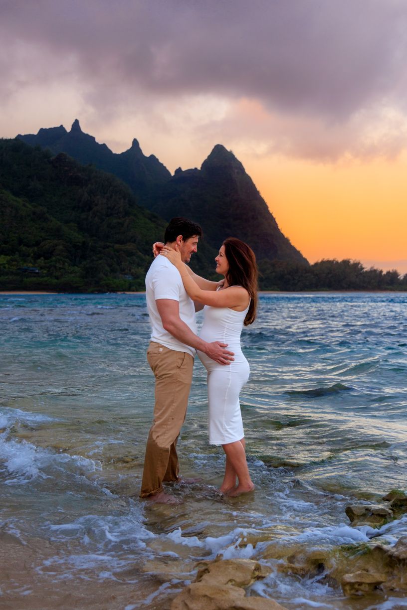 A couple wades in shallow ocean water during sunset with mountains creating a dramatic backdrop.