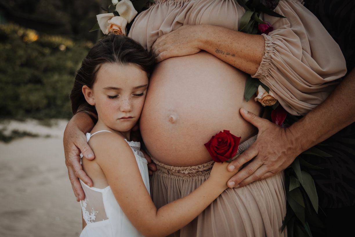 A maternity photoshoot featuring a flowing white dress and floral accents against a dark background.