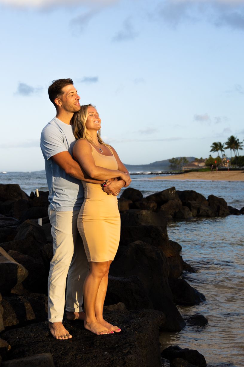 A couple stands together on rocky coastal formations during golden hour by the ocean.