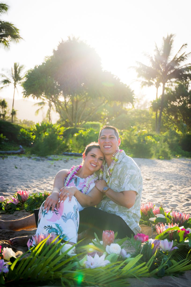 Two people lay on a sandy beach with tropical flowers and palm trees during golden hour.
