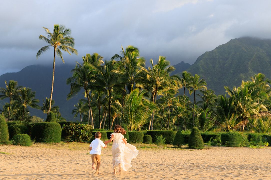 Couple running on a sandy Hawaiian beach with lush palm trees and dramatic mountain backdrop at golden hour.