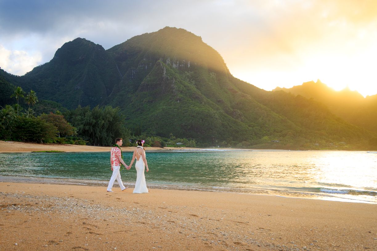 Couple walking along a scenic Hawaiian beach at sunset with mountains in the background.