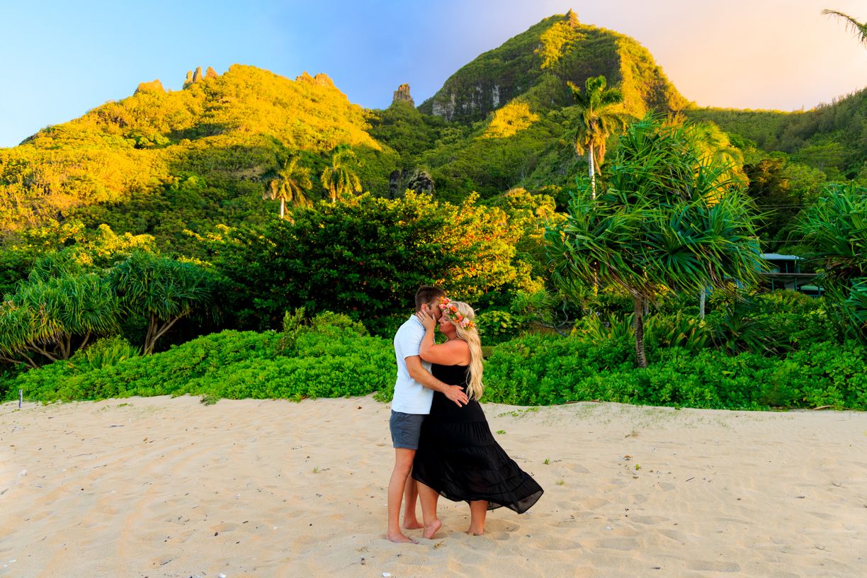 Two people share a tender moment on a sandy beach with lush mountains in the background.