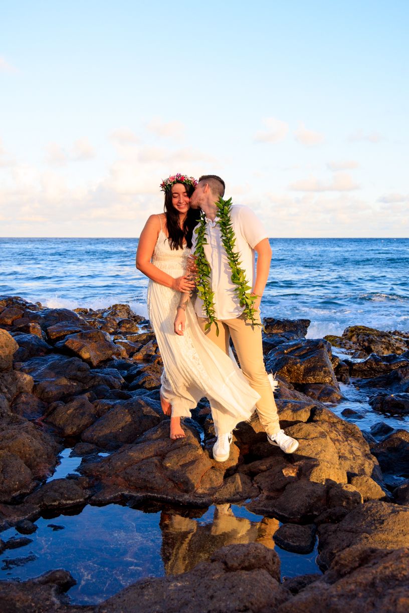A couple in wedding attire standing on volcanic rocks by the ocean at sunset in Hawaii.