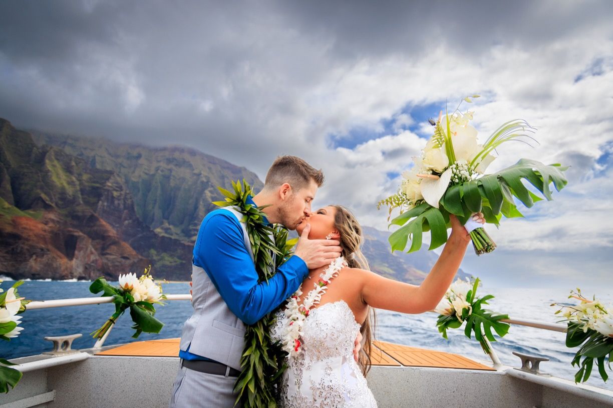 Newlyweds sharing romantic moment on boat with tropical flowers and coastal cliffs behind them.