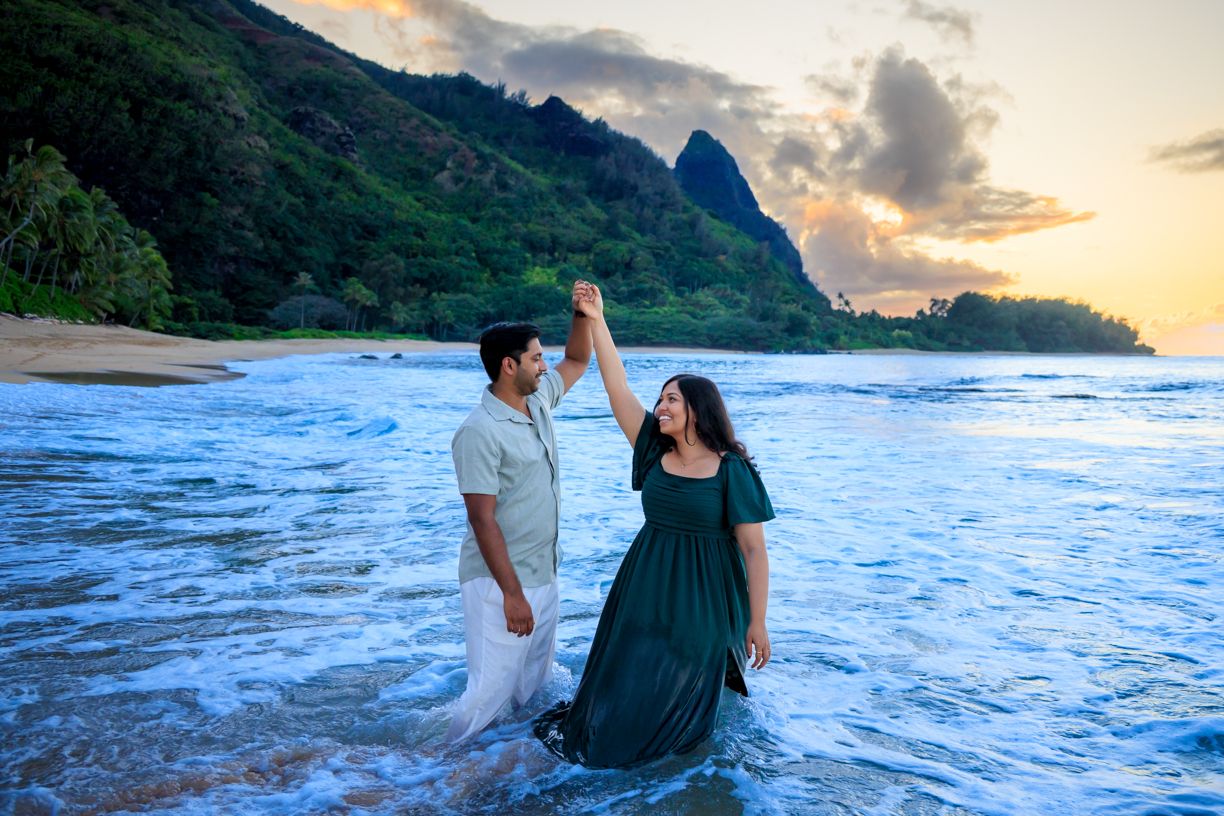 A couple holds hands while dancing in ocean waves with mountains and sunset in the background.
