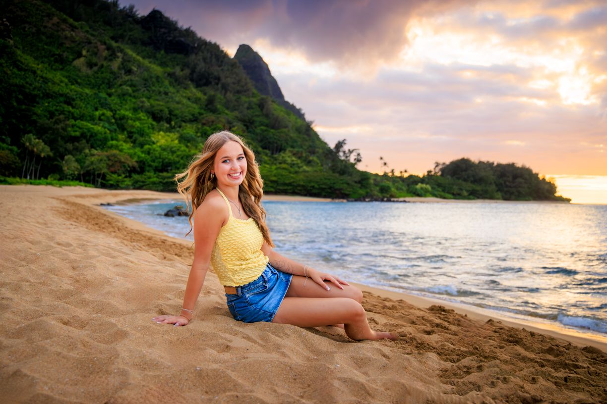 Woman in yellow top and denim shorts sitting on beach with ocean and mountains at sunset.