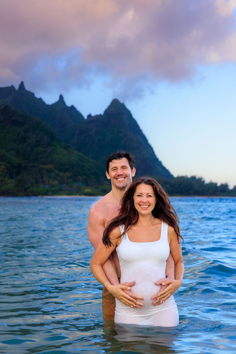 A couple shares a joyful moment in ocean waters with dramatic mountain peaks in the background.
