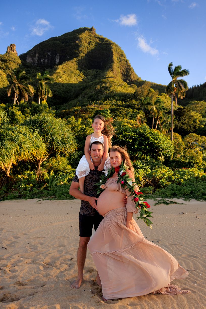 Playful maternity photos on tropical beach with mountains and palm trees at sunset.