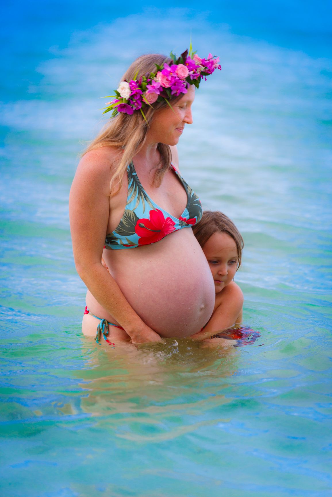Maternity photoshoot in turquoise waters with vibrant flower crown and flowing dress.