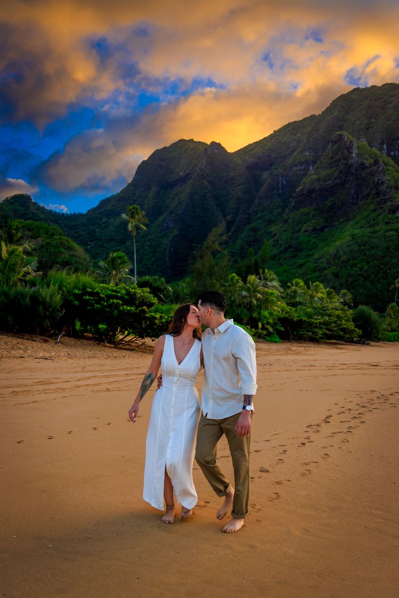 Couple walking hand in hand on a sandy beach with dramatic mountain landscape at sunset.