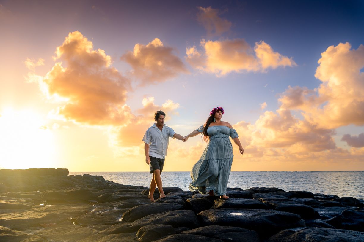 Couple holds hands on volcanic rocks at sunset with dramatic golden clouds over the ocean in Hawaii.
