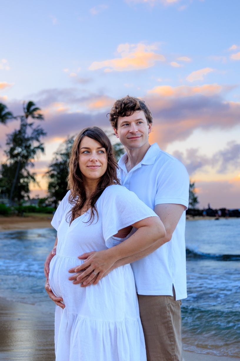 A beachside maternity photoshoot features a couple in white clothing during golden hour.
