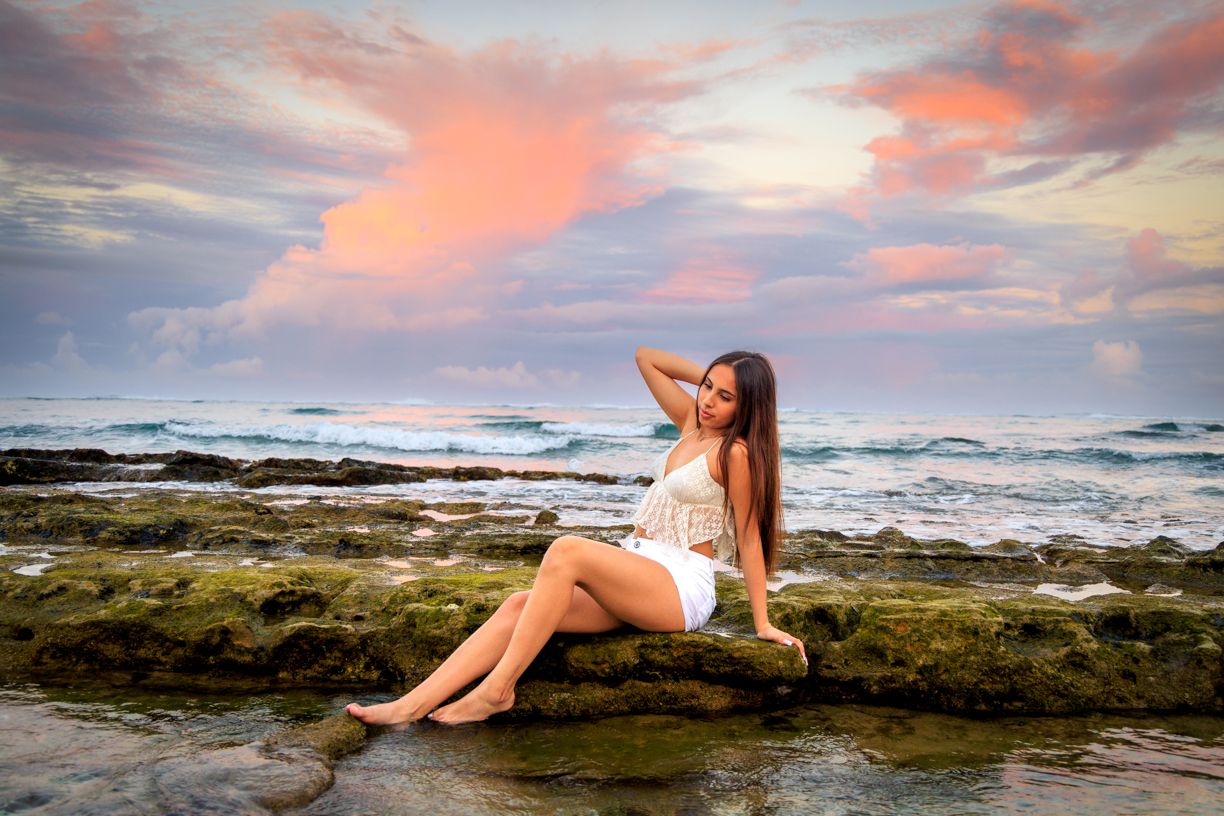 Woman in white dress sitting on rocky shore at sunset.