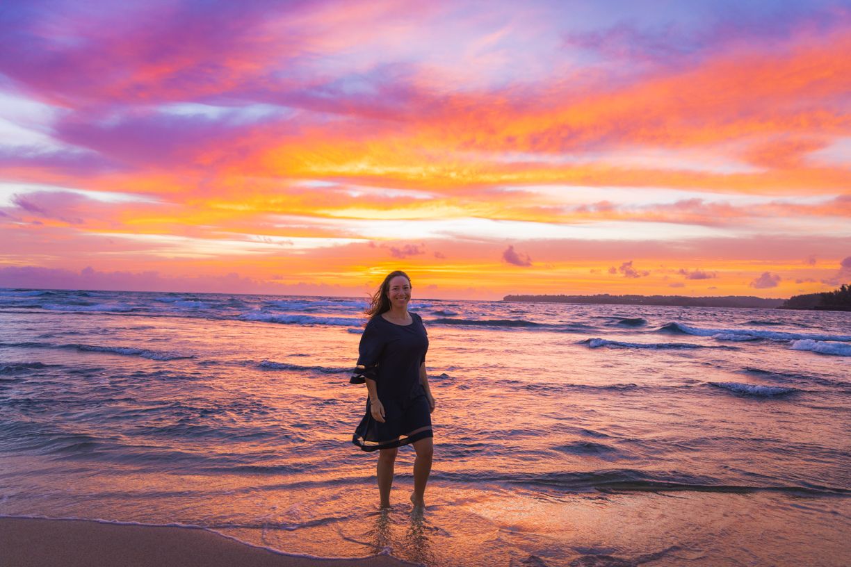 Silhouette stands in ocean waves during vibrant orange and purple sunset.