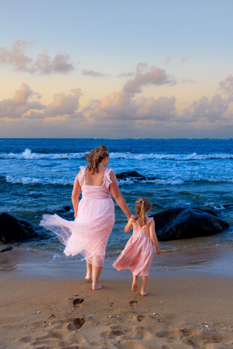 Two people in pink dresses walk along a beach at sunset with waves crashing against rocks.