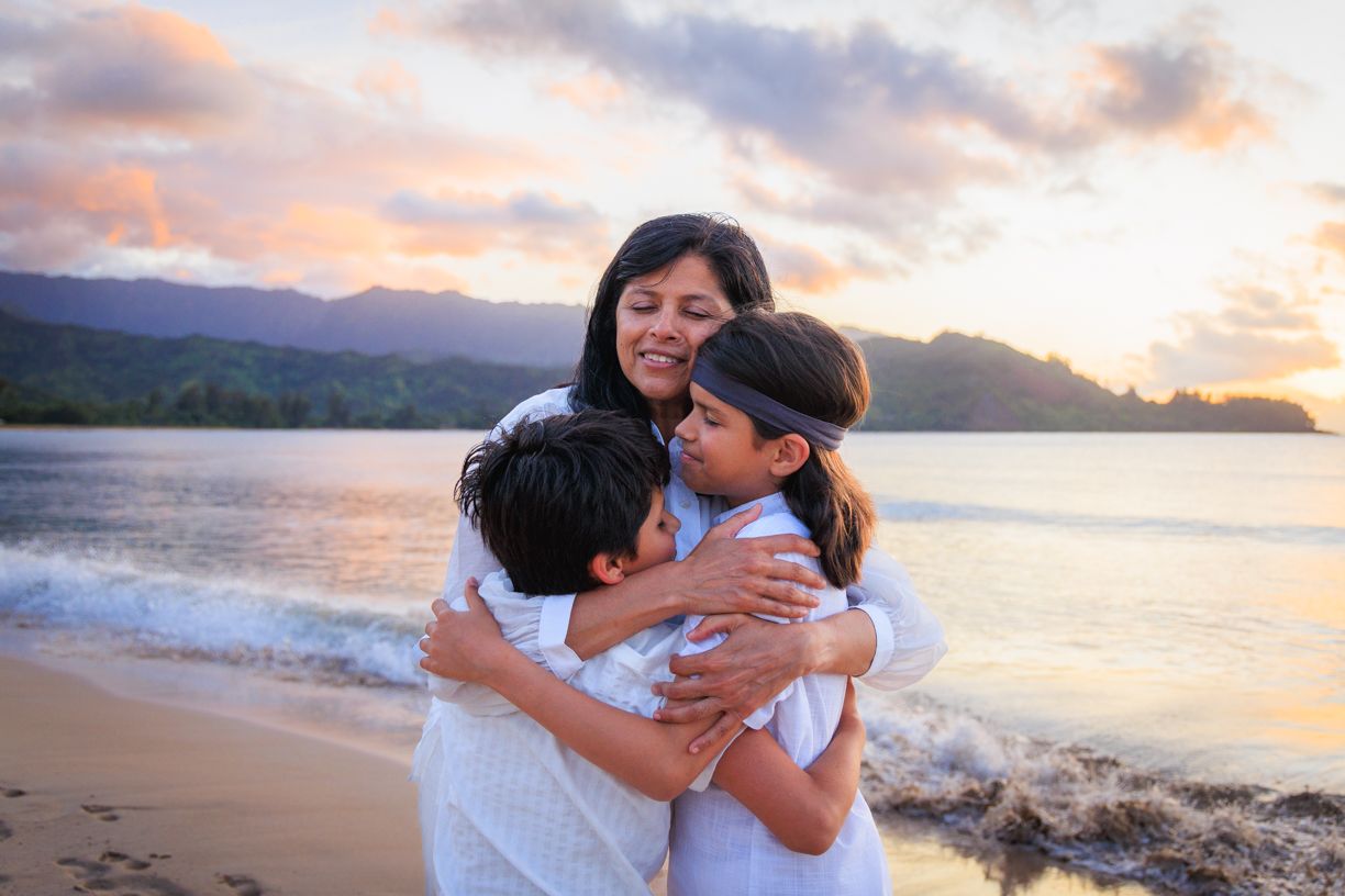 Three people hugging on beach at sunset with mountains in background.