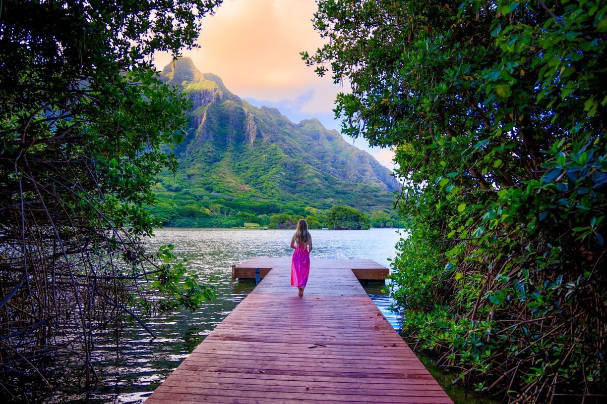 Wooden pier extends into tropical lake with lush mountains and sunset sky in background.