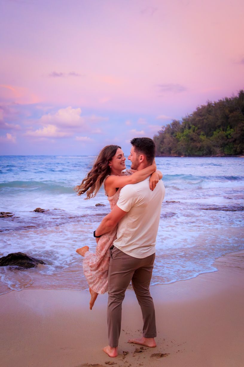 Playful couple embracing on beach during purple sunset with waves crashing nearby.