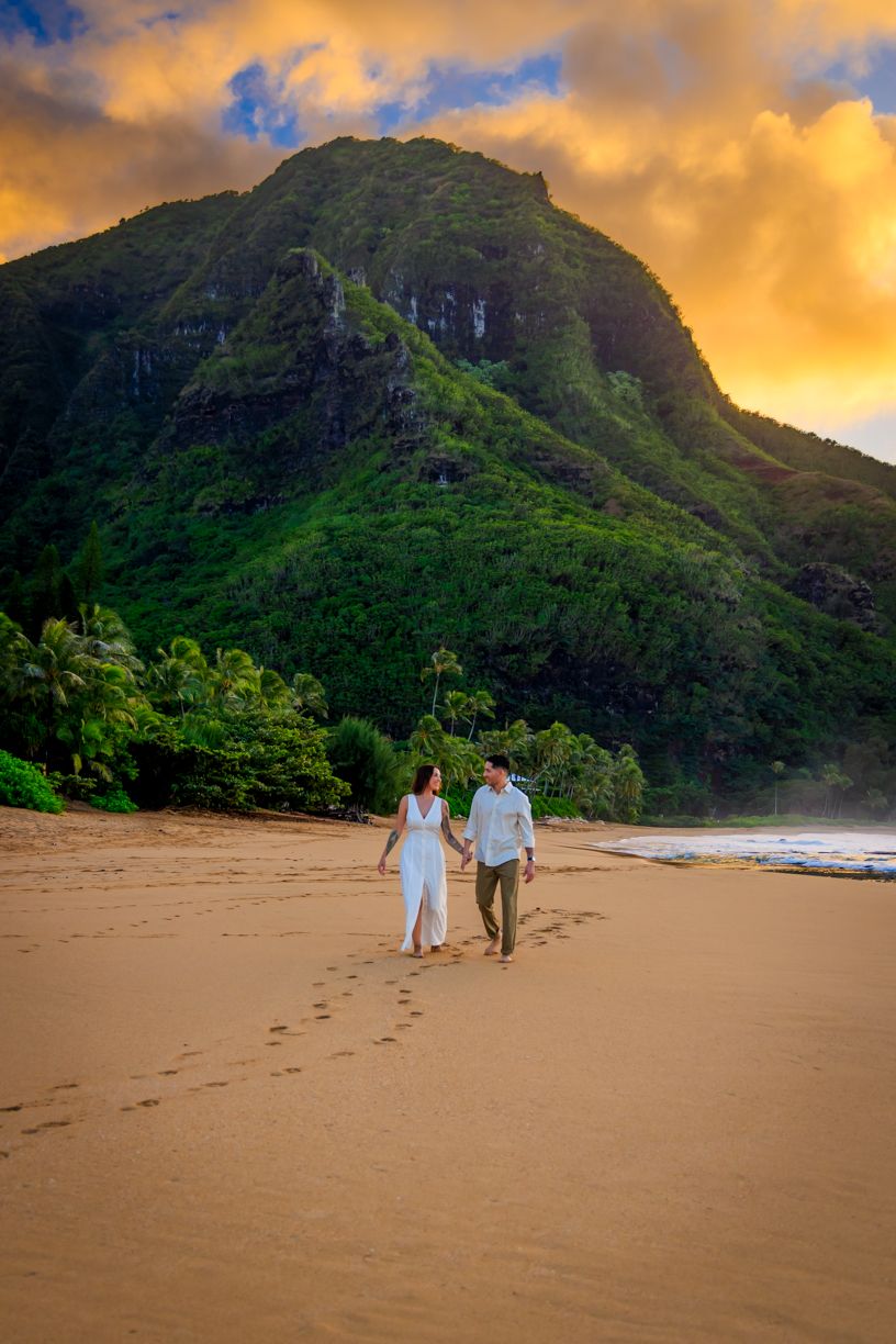 Couple walking on sandy beach with dramatic mountain cliffs and golden sunset.