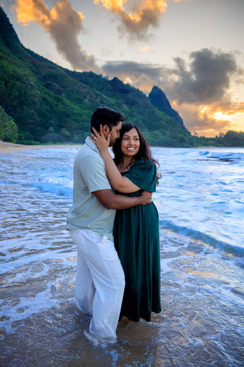 A couple embraces in the ocean waves during sunset at a Hawaiian beach.