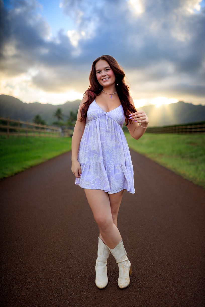 Figure in a white flowy dress and boots poses on a rural road at sunset with dramatic clouds and mountains.
