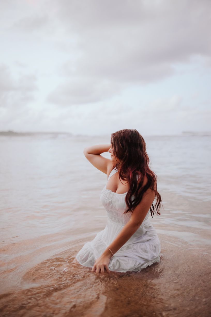 Woman in white dress sitting in shallow ocean water at sunset.
