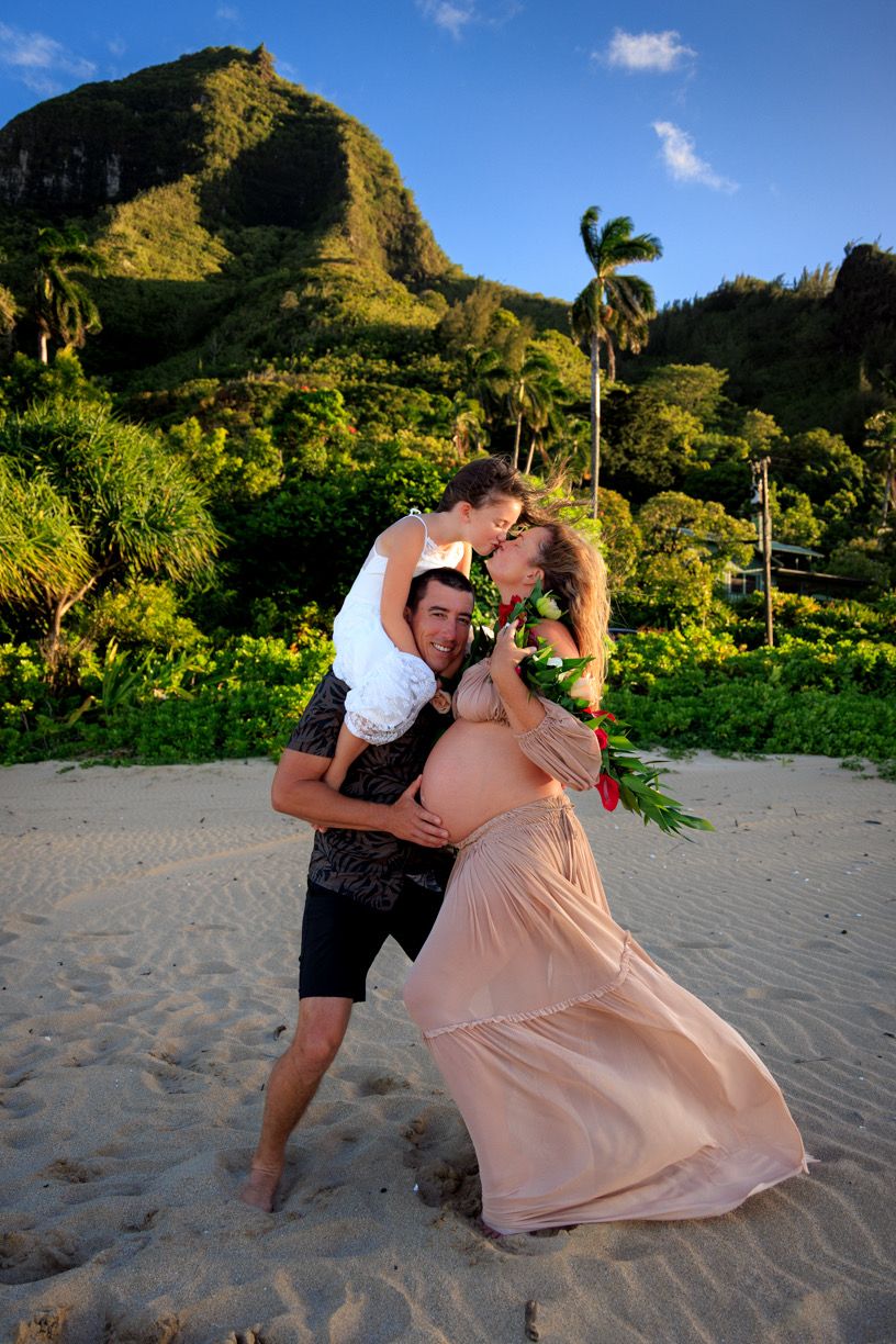 Playful maternity photography on a Hawaiian beach with mountains and palm trees.