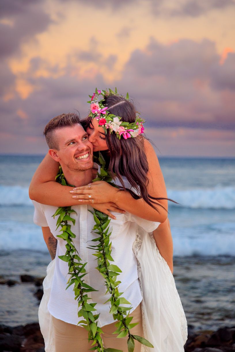 A joyful sunset beach moment with traditional Hawaiian leis and flower crown against ocean waves.
