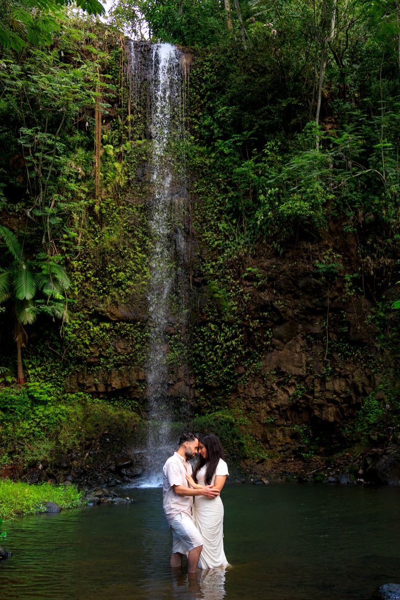 A romantic waterfall photoshoot in Hawaii with a couple standing in a natural pool surrounded by jungle.