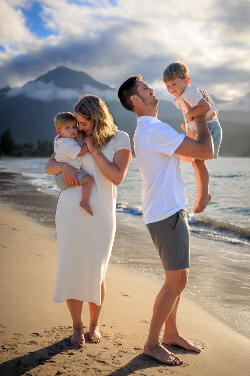 Family of four enjoying a sunset beach day, parents holding young children with mountains and ocean in the background.