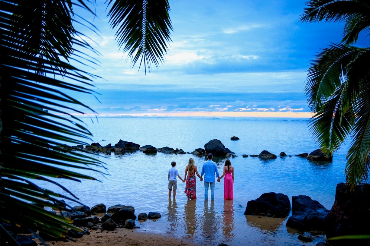 Family holding hands on a tropical beach at sunset, framed by palm trees with rocky shoreline and vibrant blue sky.