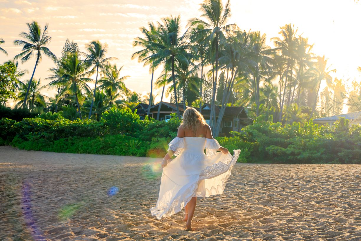 Woman in white dress dancing on sandy beach at sunset with tropical palm trees in golden light.