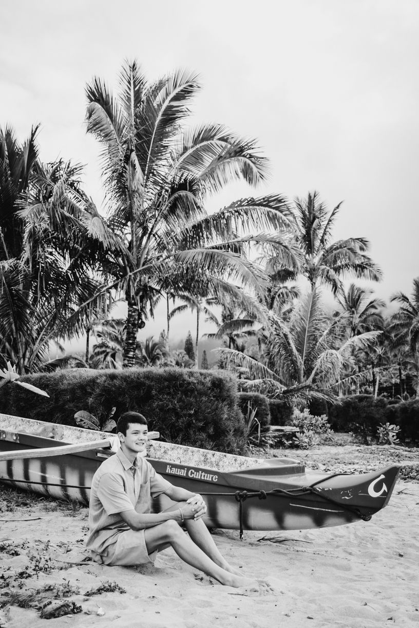 Vintage black and white photo of someone sitting beside an outrigger canoe on a tropical beach.