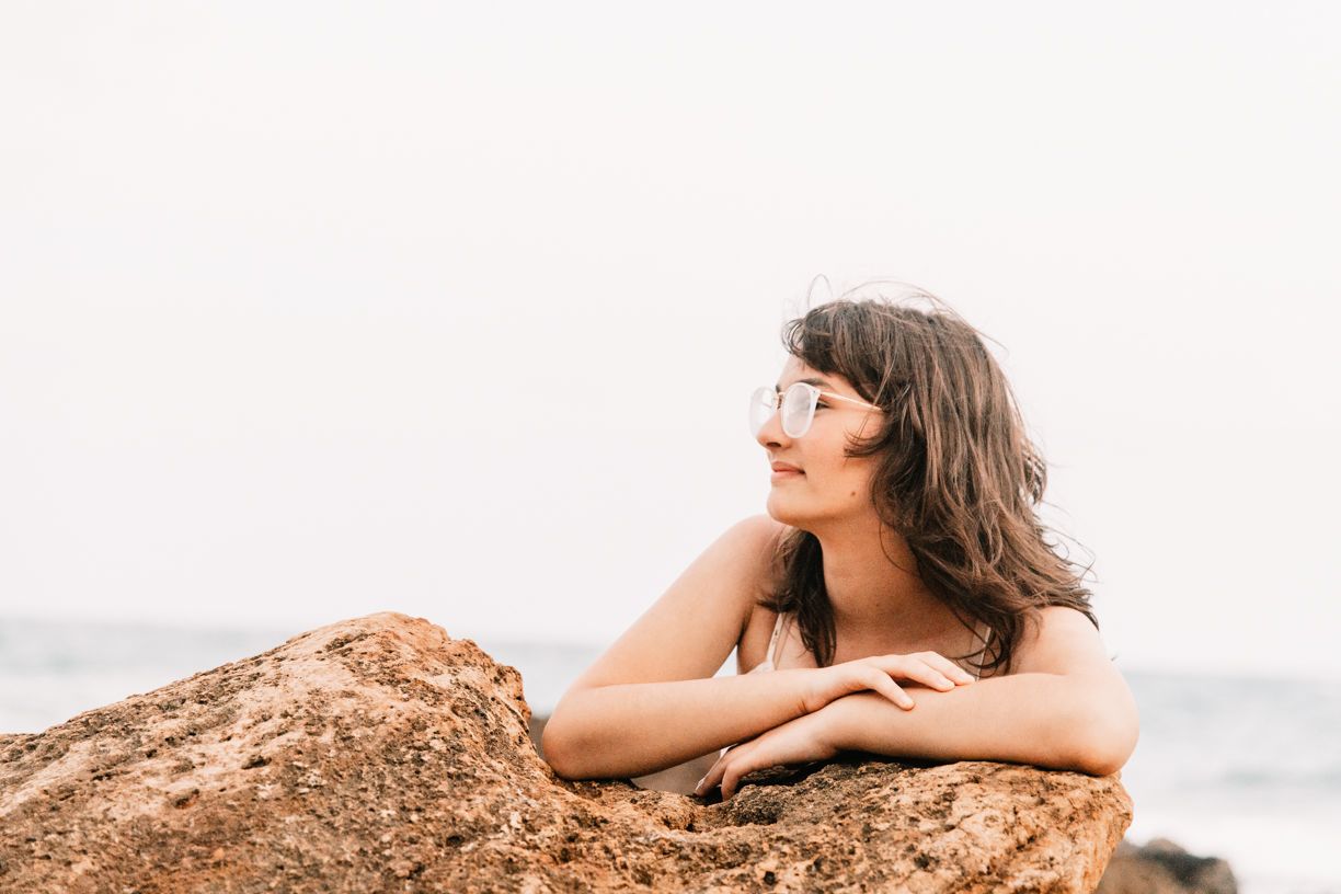 Woman with sunglasses resting on a large rock by the ocean.
