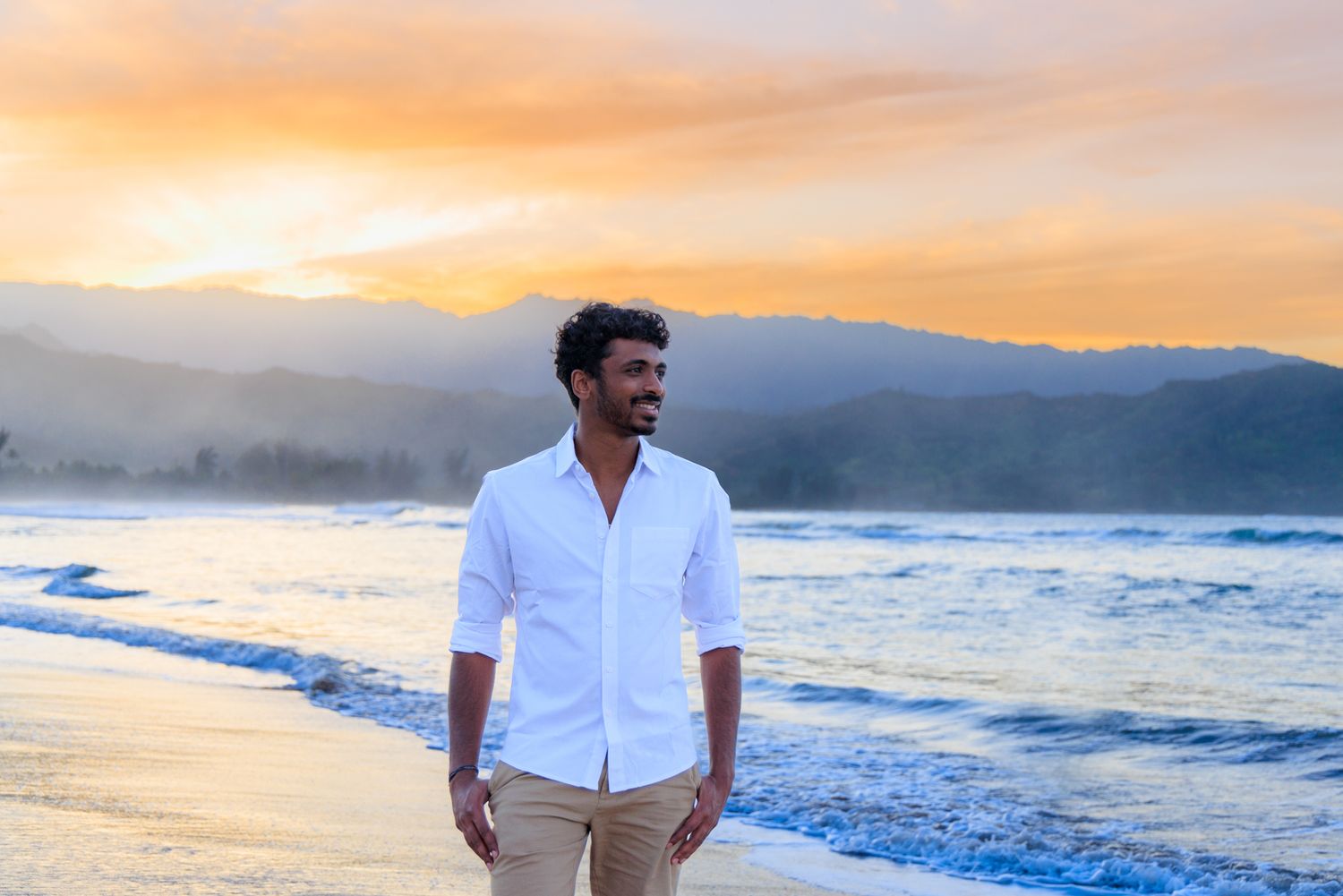 Man in white shirt smiling on beach at sunset with mountains and ocean waves in background.