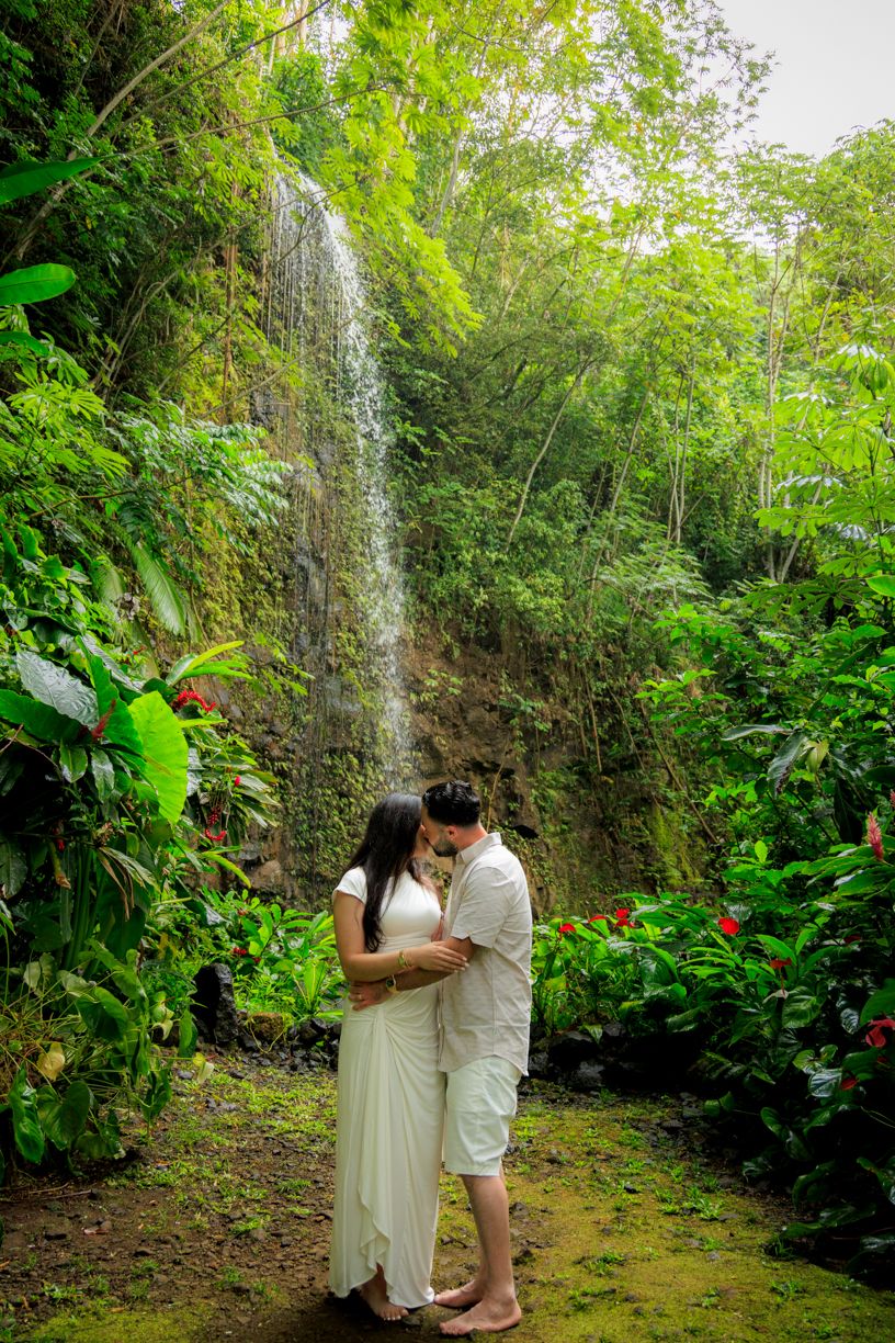 A couple shares an intimate moment by a tropical waterfall surrounded by lush greenery.