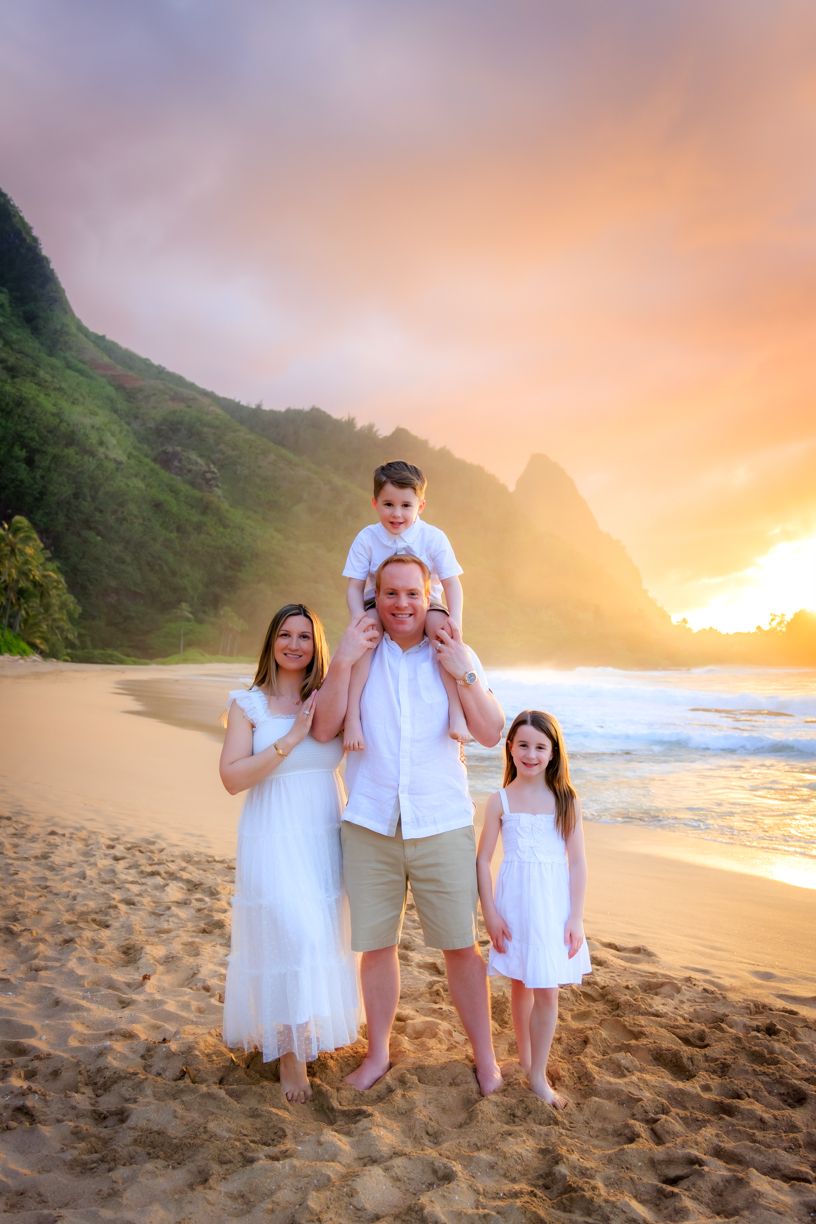 Family of four posing on a sandy Hawaiian beach at golden sunset with lush green mountains in the background.