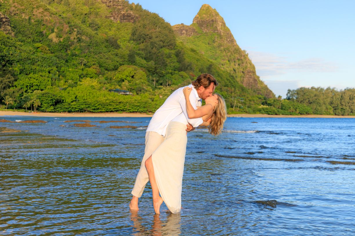 A couple wades in crystal clear tropical waters with mountains visible in the distance.