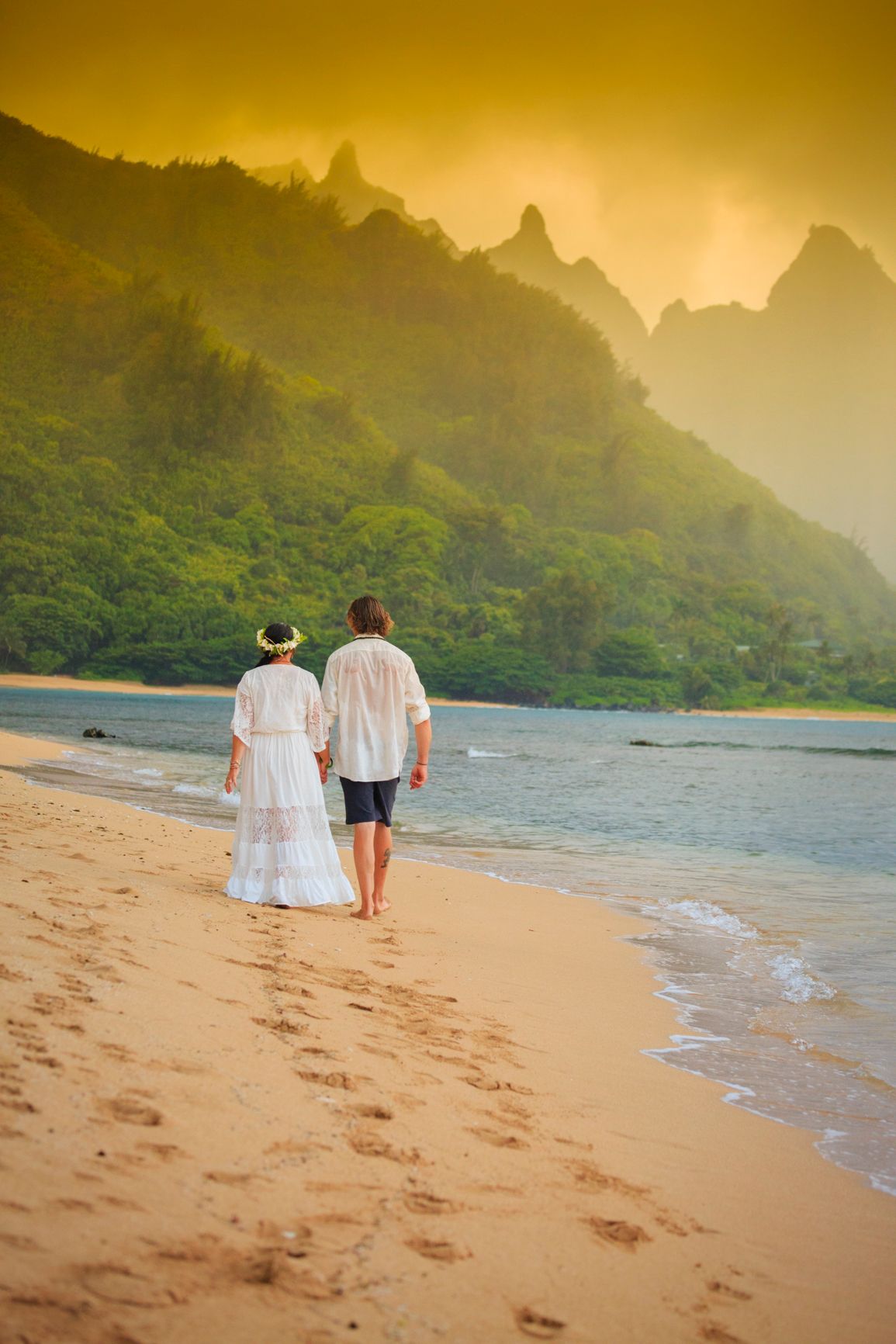 Couple walking along golden sunset beach with dramatic Na Pali coast mountains in background.