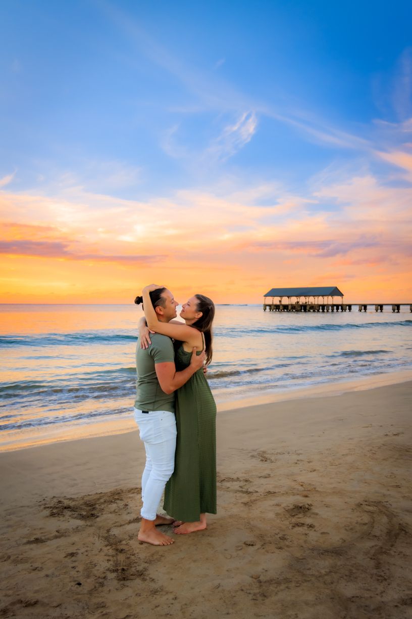 Loving couple embracing on beach with pier and sunset backdrop.