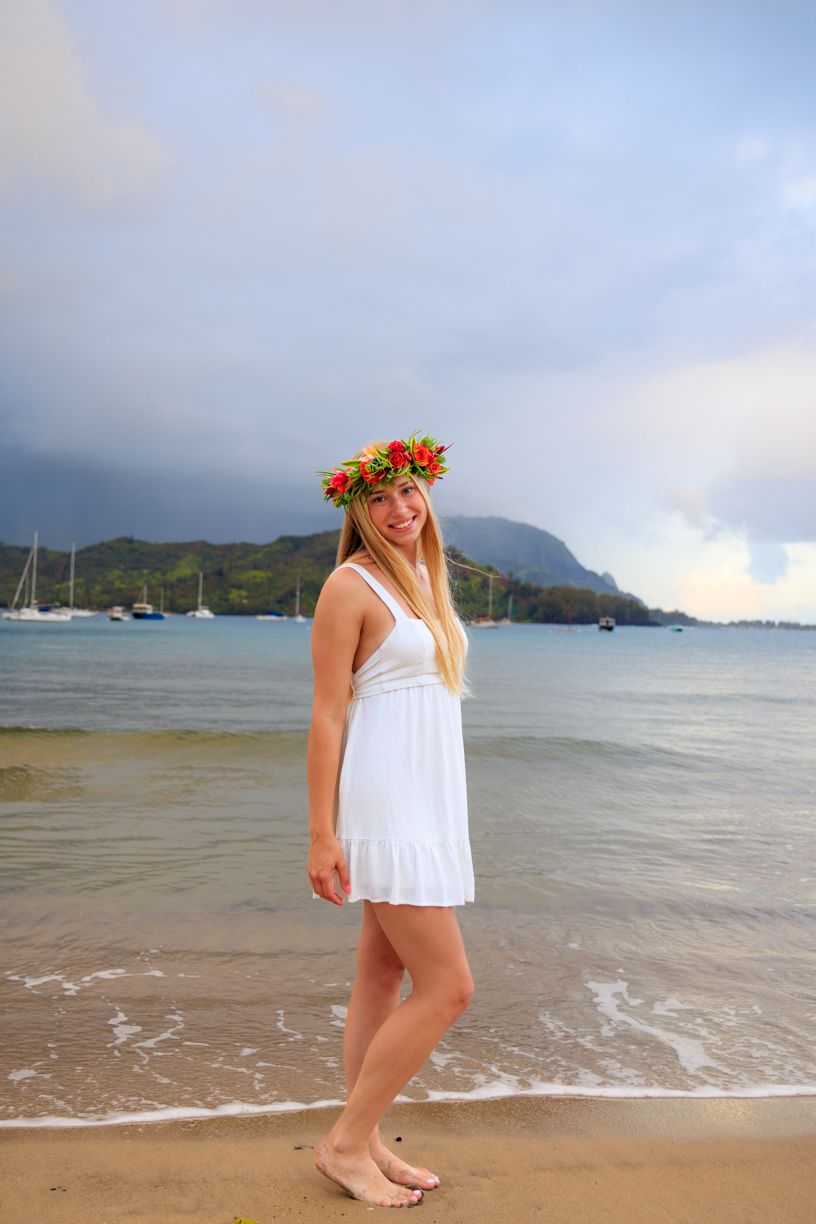 Woman in white dress and flower crown standing on beach with mountains in background.