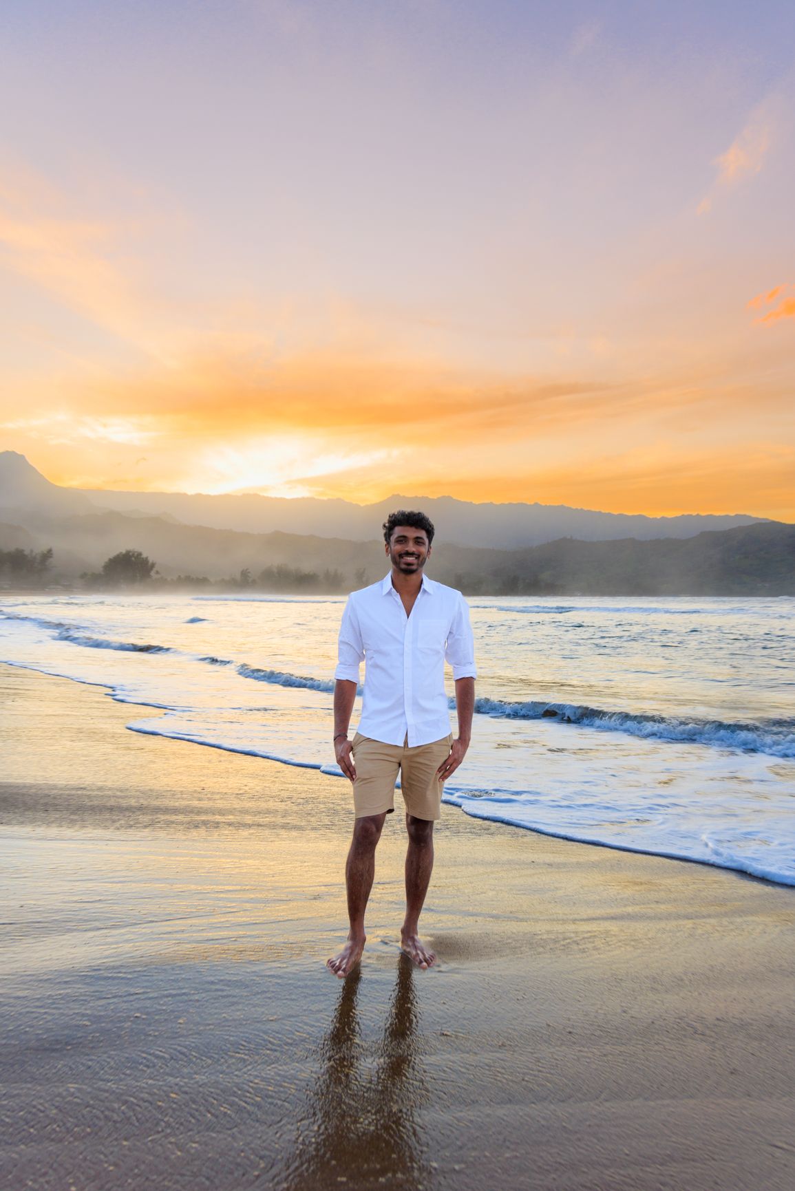 Man in white shirt and khaki shorts standing on beach at sunset with mountains in background.