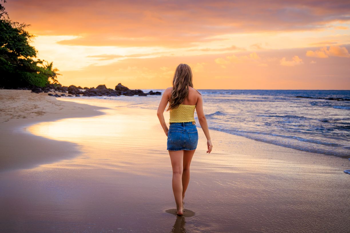 Woman walking on beach at sunset with ocean and mountains in background.