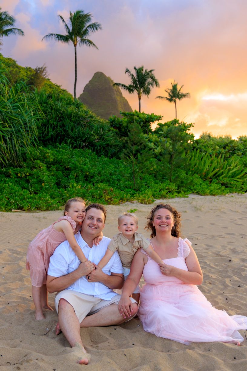 A family sits together on a sandy beach with tropical mountains and palm trees at sunset.