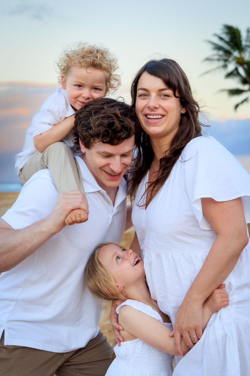 Family in white clothing sharing playful moments on beach at sunset.