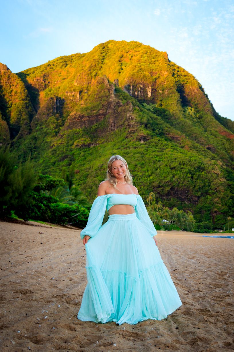 A turquoise dress billows in the wind on a Hawaiian beach with dramatic mountain peaks in the background.